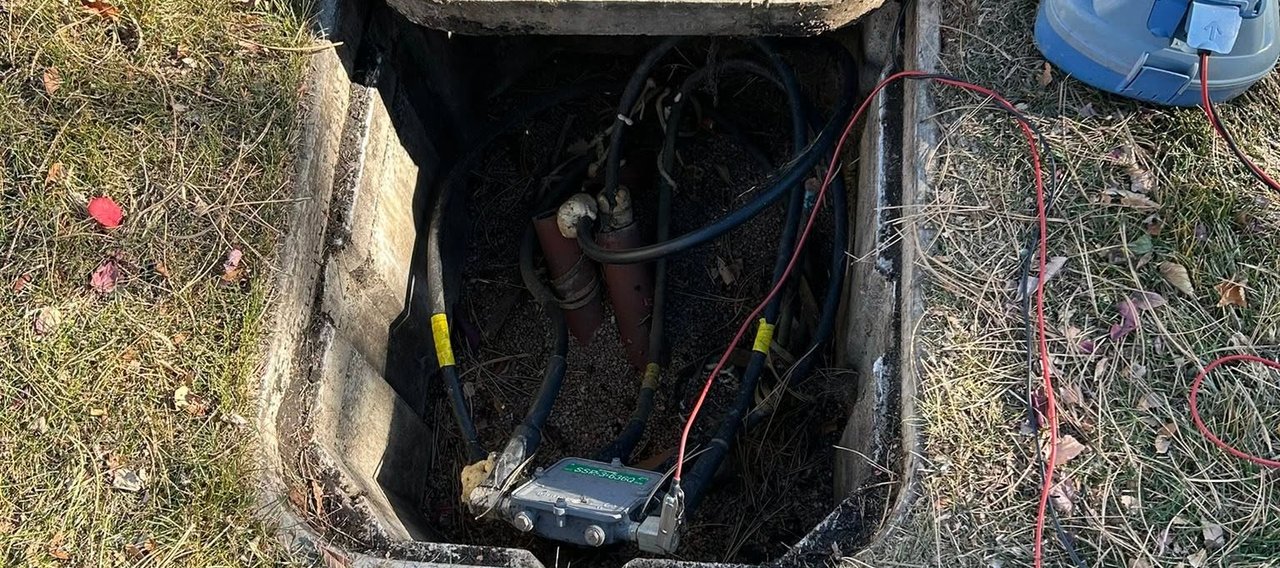 Open Comcast underground utility vault with cables and a splice box, connected to a handheld test meter on the grass