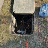 Open Comcast underground utility vault with cables and a splice box, connected to a handheld test meter on the grass
